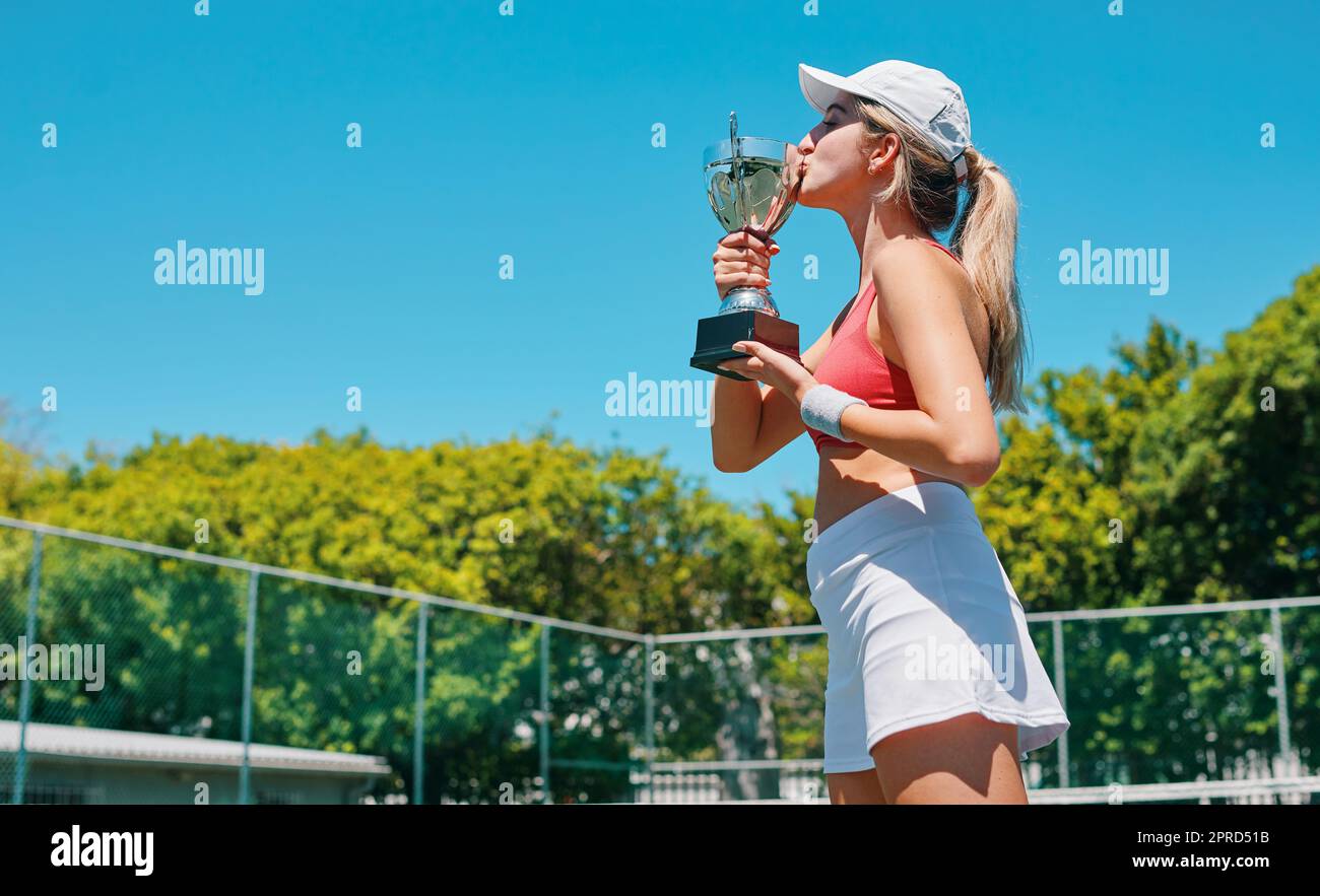 Ein weiterer Sieg unter meinem Gürtel. Eine attraktive junge Frau steht und küsst ihre Trophäe, nachdem sie tagsüber ein Tennismatch gewonnen hat. Stockfoto