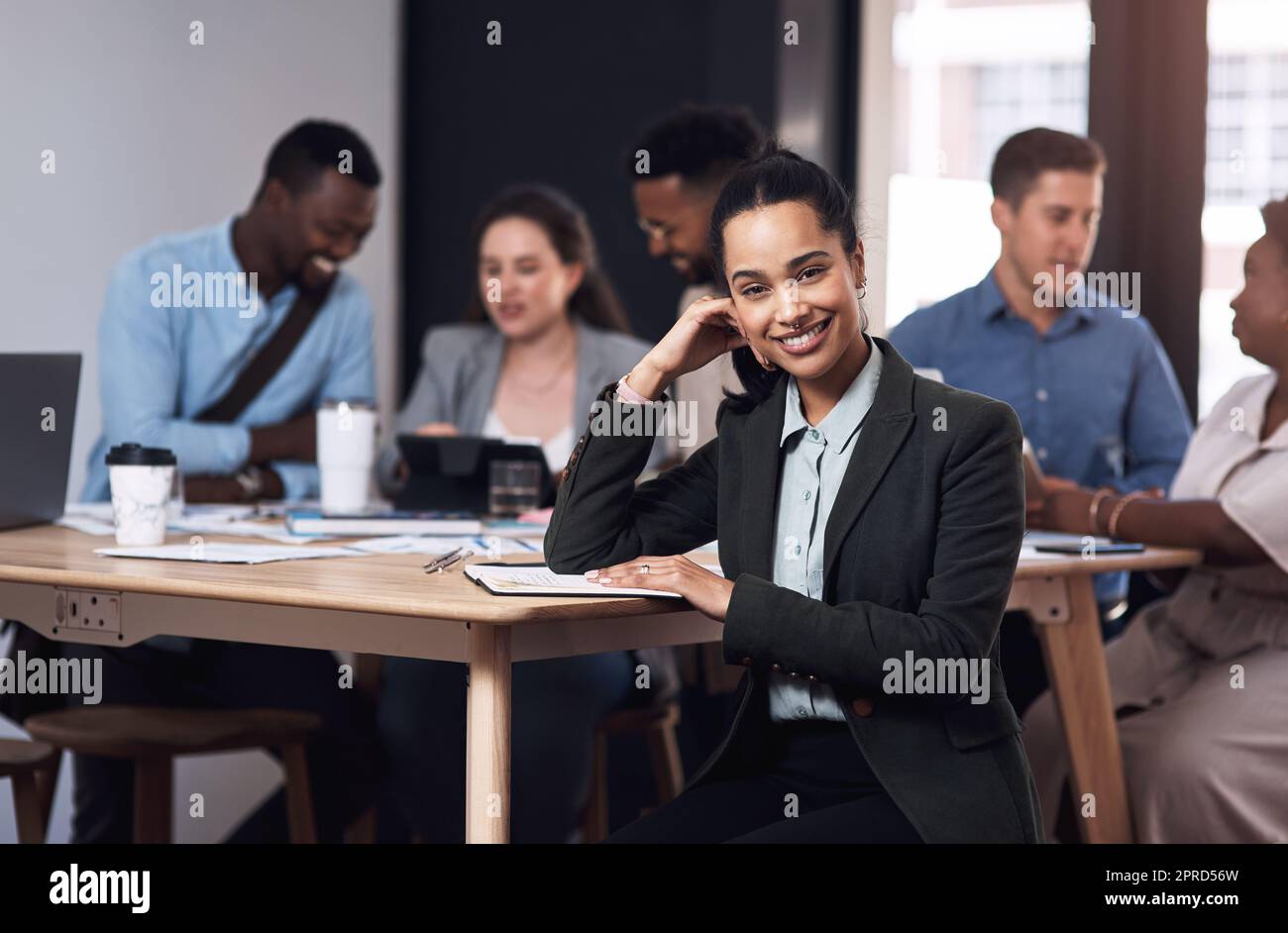 Selbstbewusst in ihrer Karriere. Porträt einer jungen Geschäftsfrau, die in einem Büro mit ihren Kollegen im Hintergrund sitzt. Stockfoto