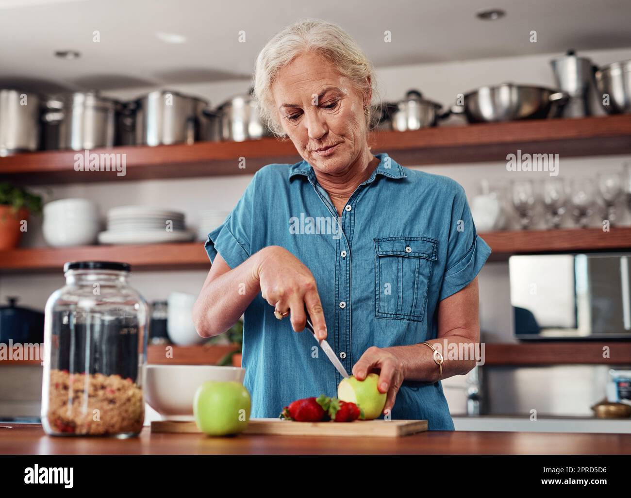 Heute isst sie nur noch gesund. Eine attraktive ältere Frau hackt Äpfel und andere Früchte, während sie das Frühstück in der Küche zubereitet. Stockfoto