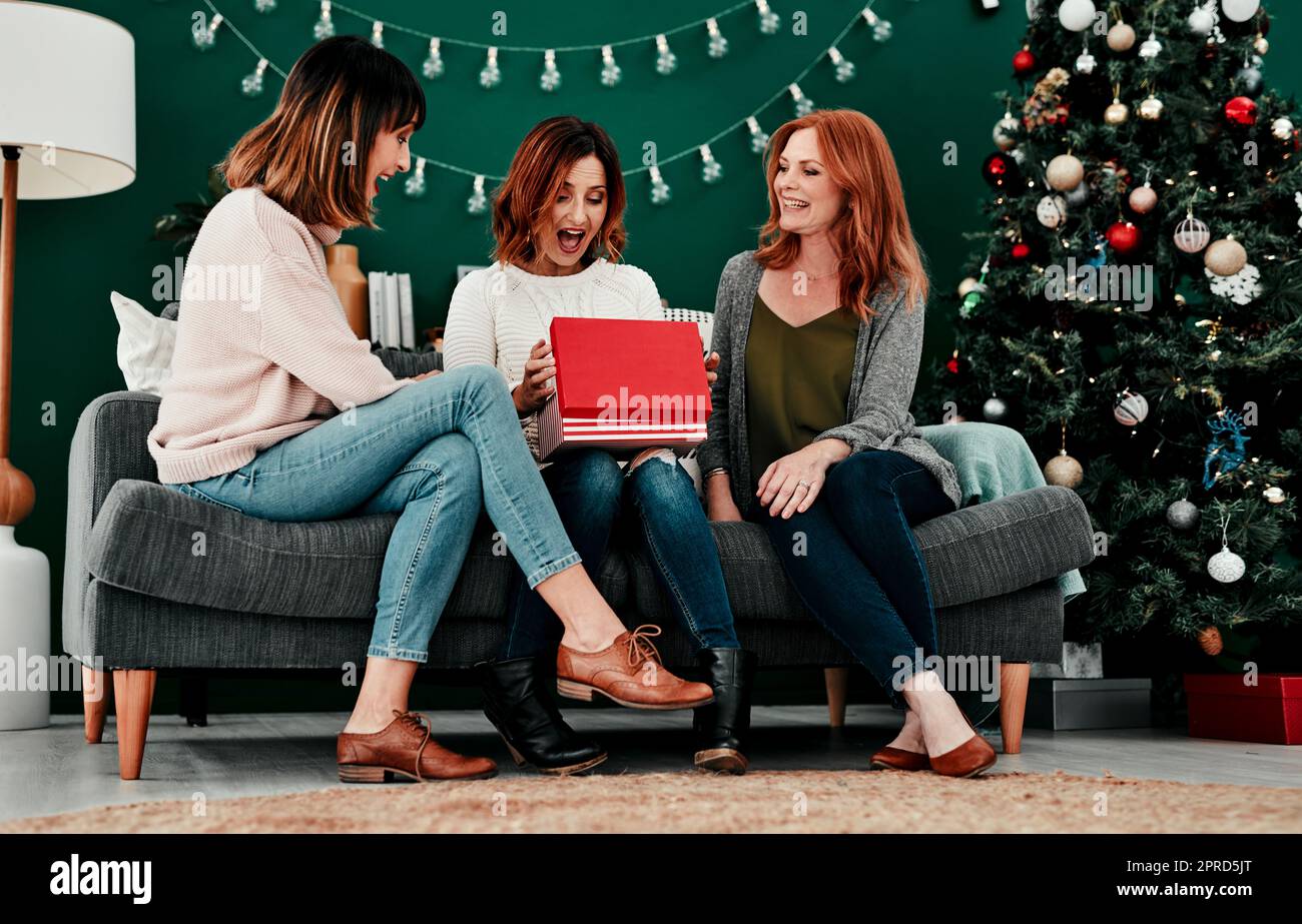 Das ist erstaunlich. Drei attraktive Frauen mittleren Alters öffnen Geschenke zusammen, während sie während der Weihnachtszeit auf einem Sofa sitzen. Stockfoto