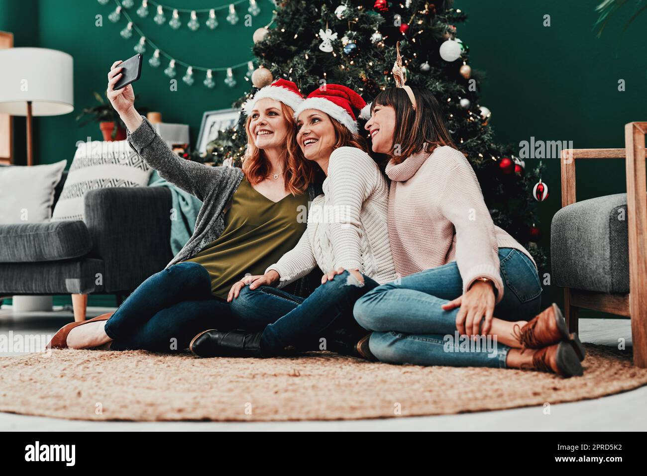 Ist jeder bereit für das Foto. Drei attraktive Frauen mittleren Alters, die während der Weihnachtszeit zusammen mit einem Handy zu Hause Selbstporträts machen. Stockfoto