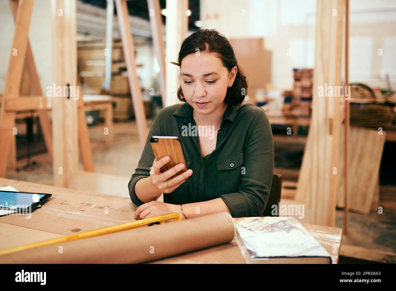 Wir bekommen so viele Kunden über Social Media. Eine Tischlerin benutzt ihr Handy, während sie in ihrer Werkstatt sitzt. Stockfoto