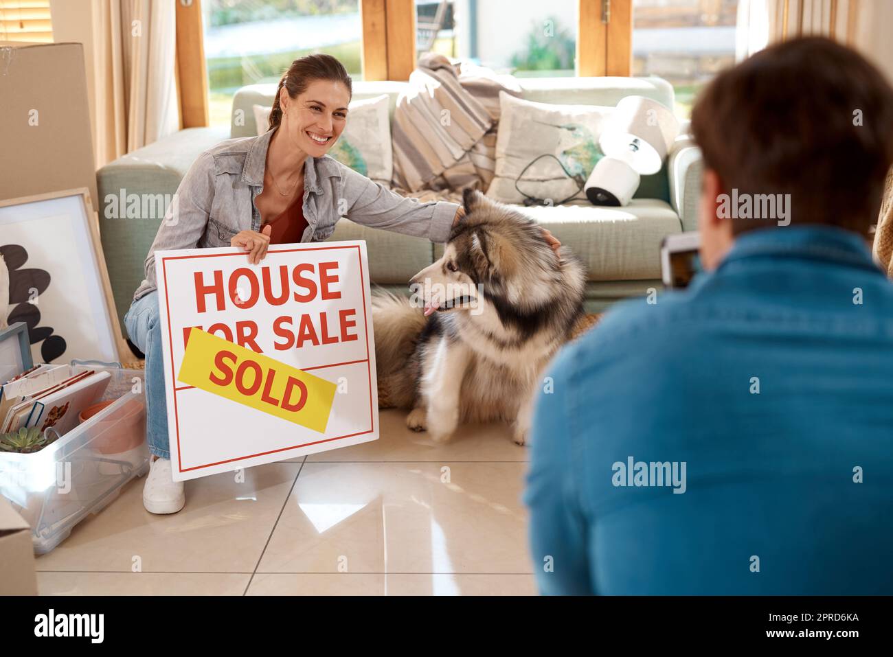 Waren die stolzen Besitzer jetzt. Ein glückliches junges Paar und ihr Hund, der Fotos mit einem verkauften Schild in ihrem neuen Zuhause macht. Stockfoto