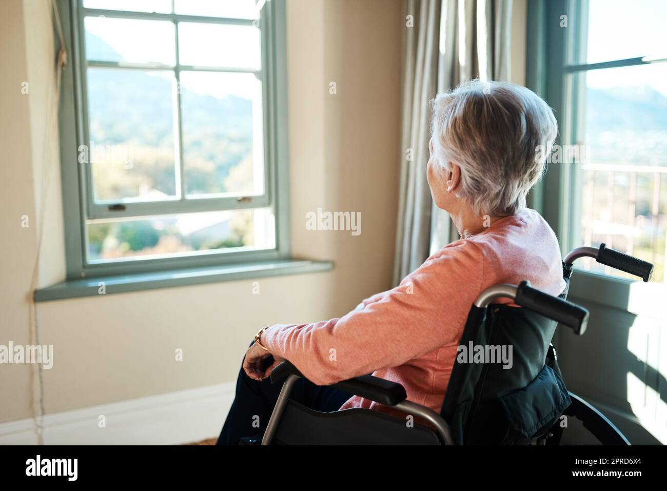 Es gibt eine ganz andere Welt da draußen. Eine ältere Frau im Rollstuhl schaut nachdenklich aus dem Fenster zu Hause. Stockfoto