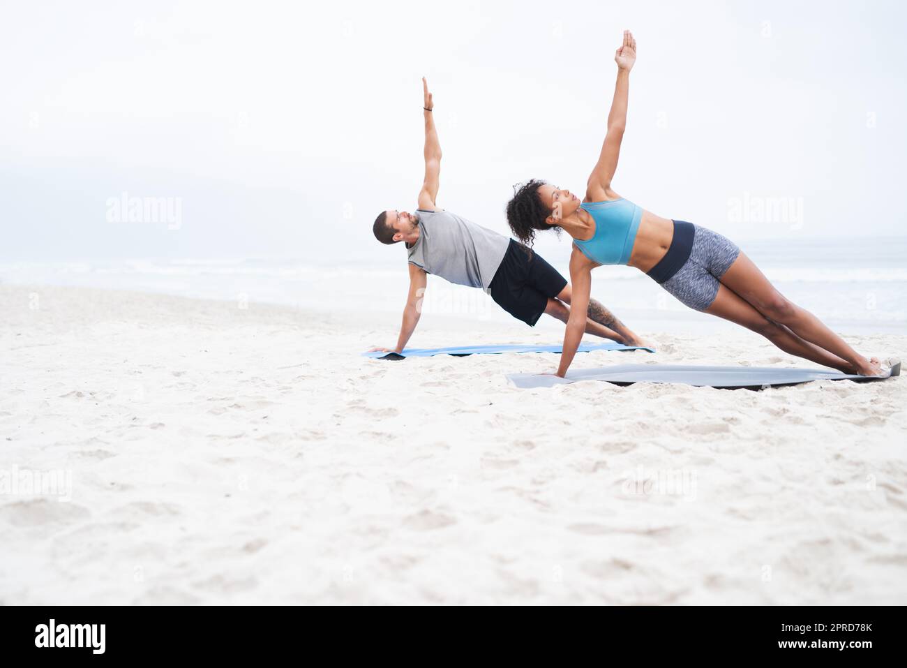 Gemeinsam einen aktiveren Lebensstil führen. Ein junger Mann und eine junge Frau üben gemeinsam am Strand Yoga. Stockfoto