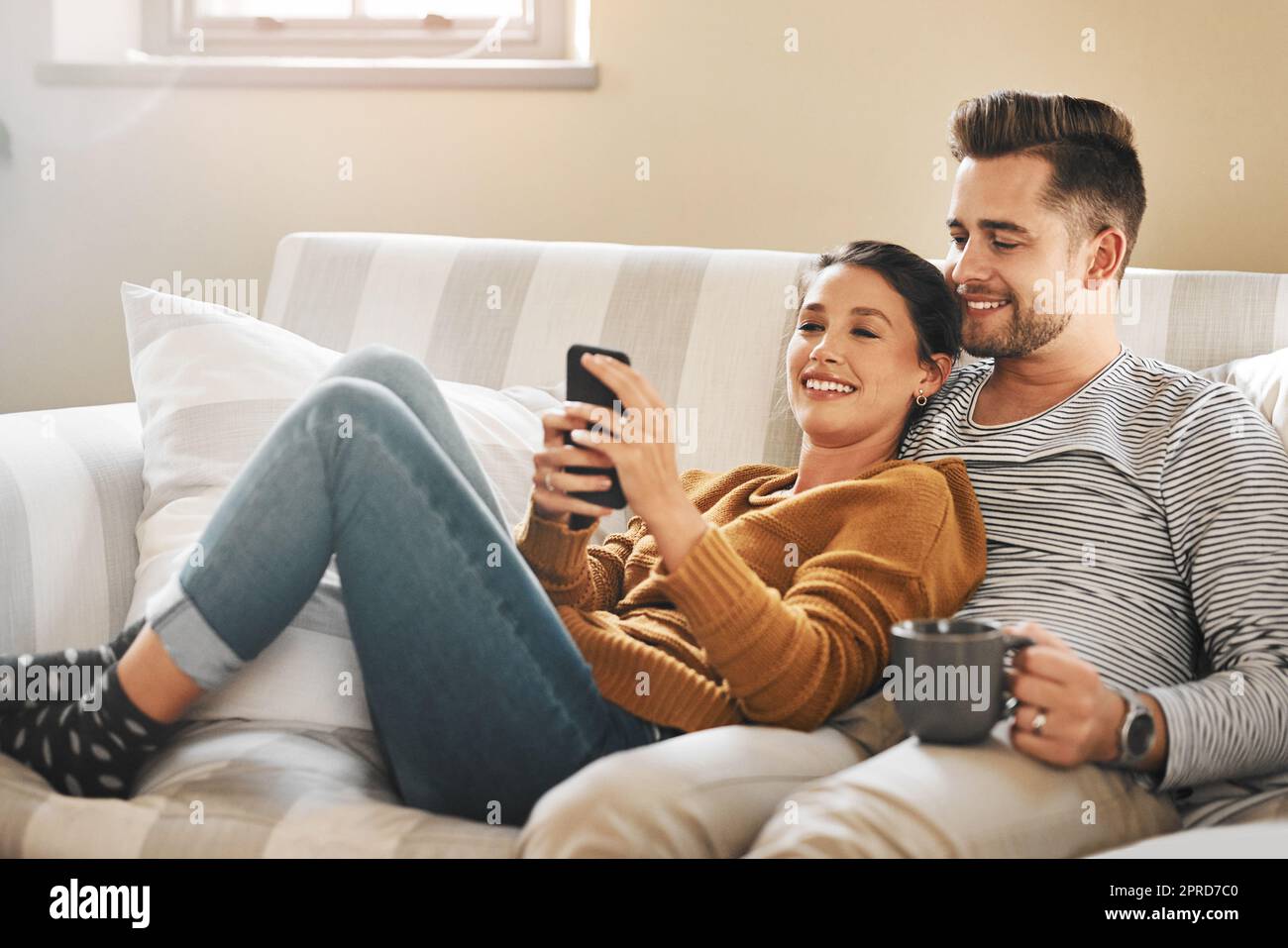 Sollten wir anrufen und hören, ob sie kommen wollen. Ein junges Paar, das ein Handy benutzt, während es sich auf dem Sofa zu Hause entspannt. Stockfoto