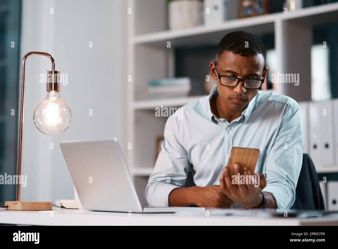 Ein hübscher junger Geschäftsmann, der seinen Laptop und sein Handy benutzt, während er spät in seinem Büro arbeitet. Stockfoto