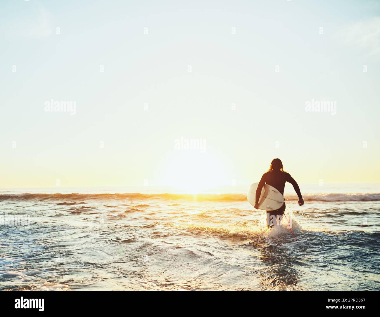 Sei gleich wieder da - ich muss ein paar Wellen fangen. Rückansicht eines jungen Mannes, der zum Meer läuft, während er ein Surfbrett am Strand trägt. Stockfoto