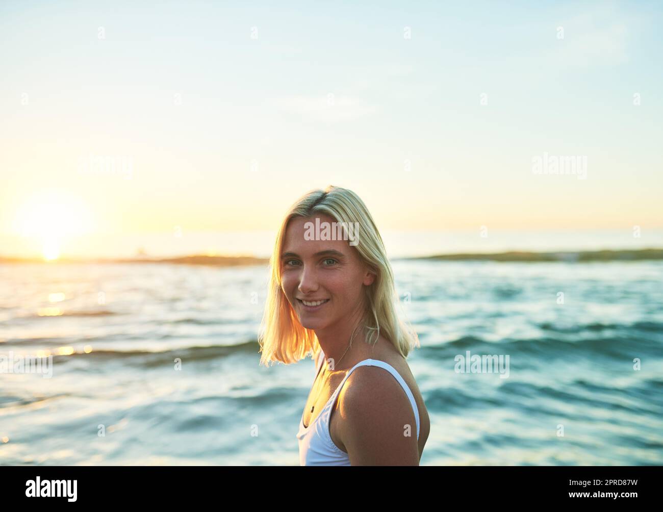 In verzweifelter Notwendigkeit von Vitamin Meer. Ein Porträt einer attraktiven jungen Frau, die bei Sonnenuntergang über ihrer Schulter am Strand lächelt. Stockfoto