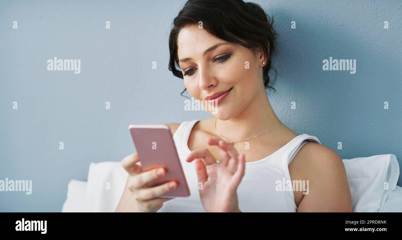 Dies wird als mein Profilbild funktionieren. Eine attraktive junge Frau, die ihr Smartphone benutzt, während sie zu Hause im Bett sitzt. Stockfoto