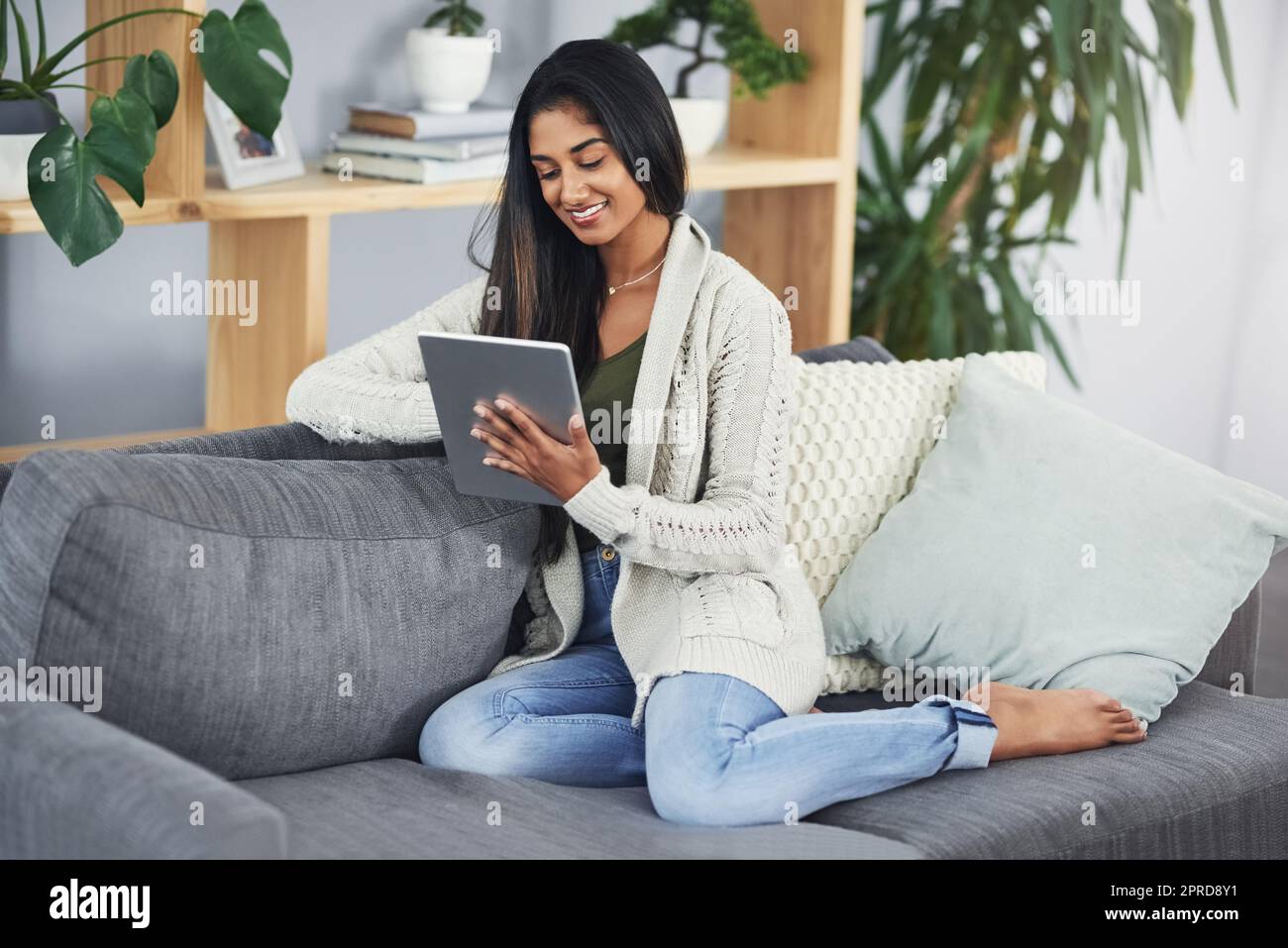 Finden Sie die beste Seite, um Ihren freien Tag zu unterhalten. Eine glückliche junge Frau mit ihrem digitalen Tablet, während sie sich auf ihrer Couch zu Hause entspannt. Stockfoto