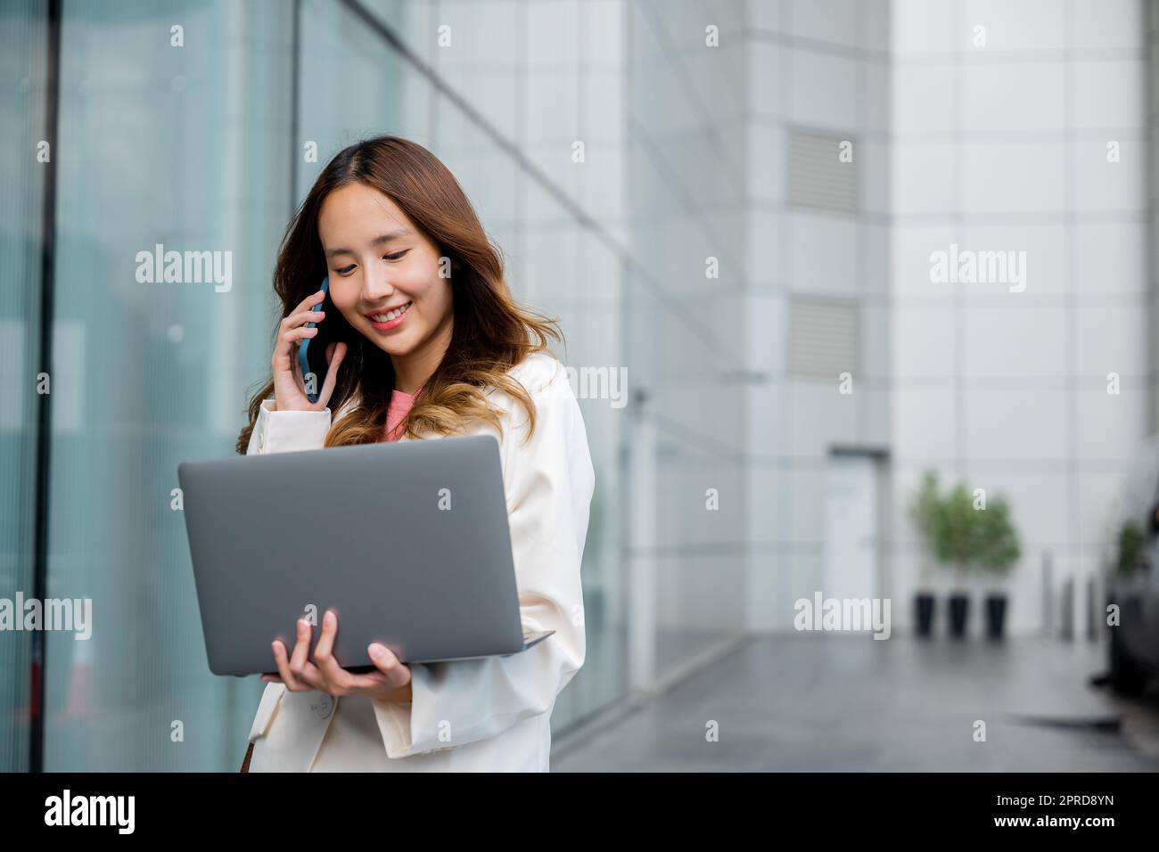 Asiatische Geschäftsfrau, die an einem Laptop arbeitet und am Handy telefoniert, im vorderen Gebäude in der Nähe des Büros Stockfoto