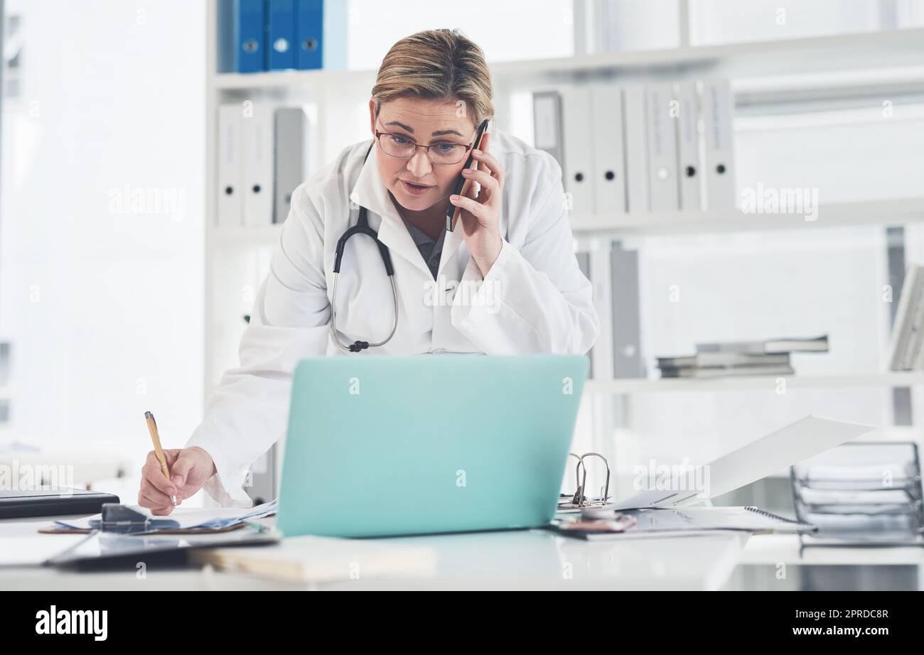 Eine attraktive junge Ärztin, die auf einem Klemmbrett schreibt, während sie in ihrem Büro einen Telefonanruf abnimmt. Stockfoto