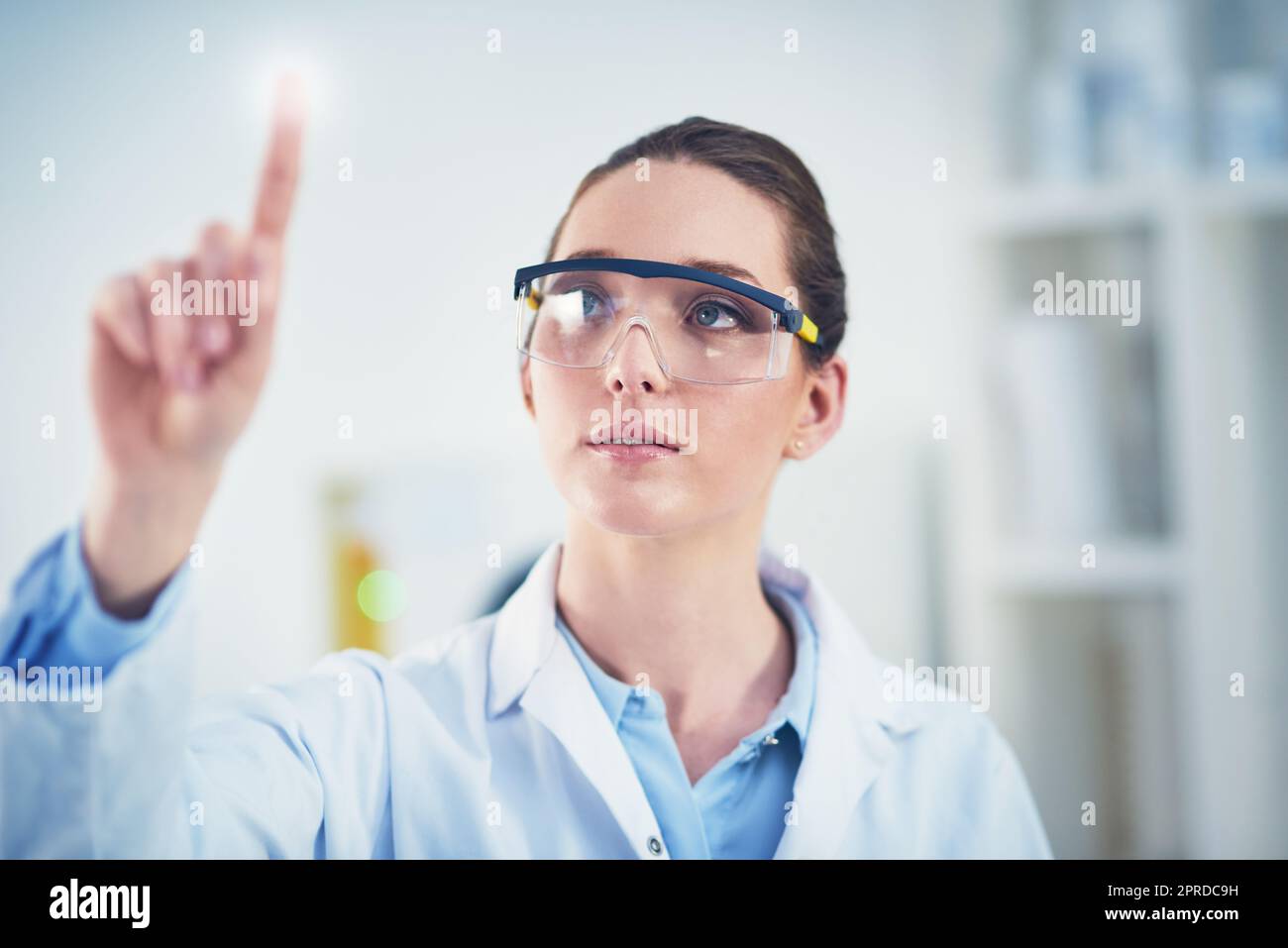 Innovation ist der Name des Spiels. Eine fokussierte junge Wissenschaftlerin trägt eine Schutzbrille und zeigt mit dem Finger auf eine Glaswand. Stockfoto