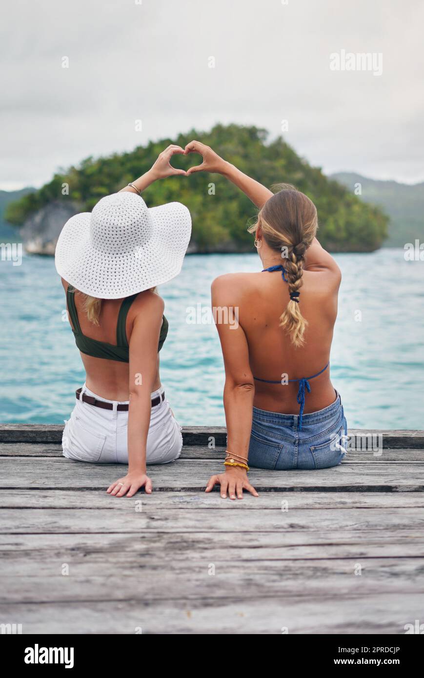 Freunde, die zusammen reisen, bleiben zusammen. Rückansicht von zwei unbekannten Frauen, die am Rande eines Boardwalk sitzen und eine Herzzeichen-Geste machen. Stockfoto