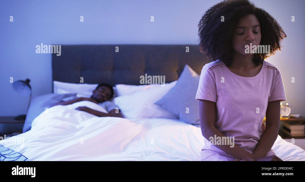 Ich habe eine große Entscheidung zu treffen. Eine besorgt aussehende junge Frau, die auf einem Bett sitzt und ihr Mann im Hintergrund schläft. Stockfoto