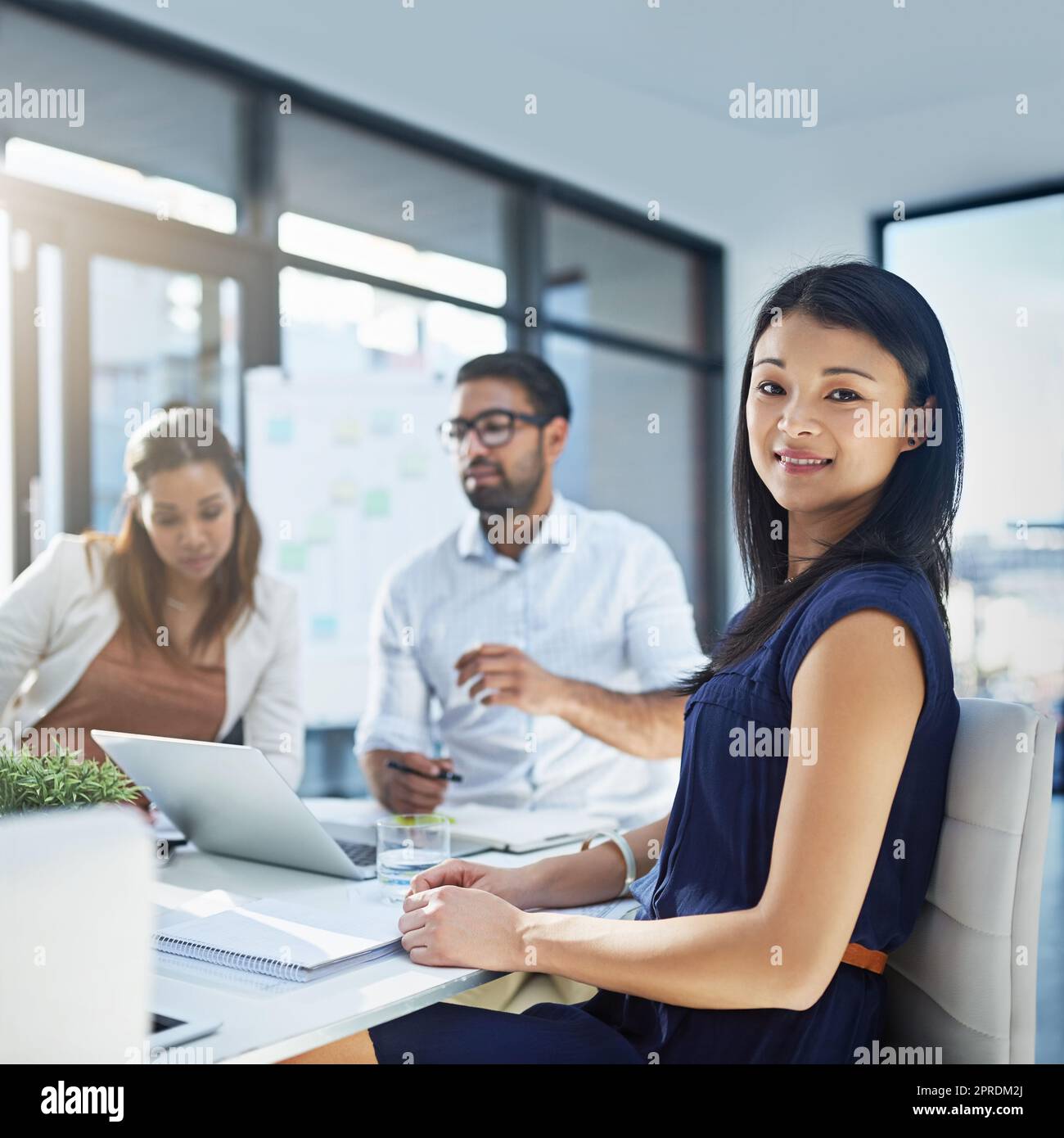 Wir bieten den besten Service der Branche. Porträt einer attraktiven jungen Geschäftsfrau, die in ihrem Büro sitzt, während ihre Kollegen im Hintergrund eine Diskussion führen. Stockfoto