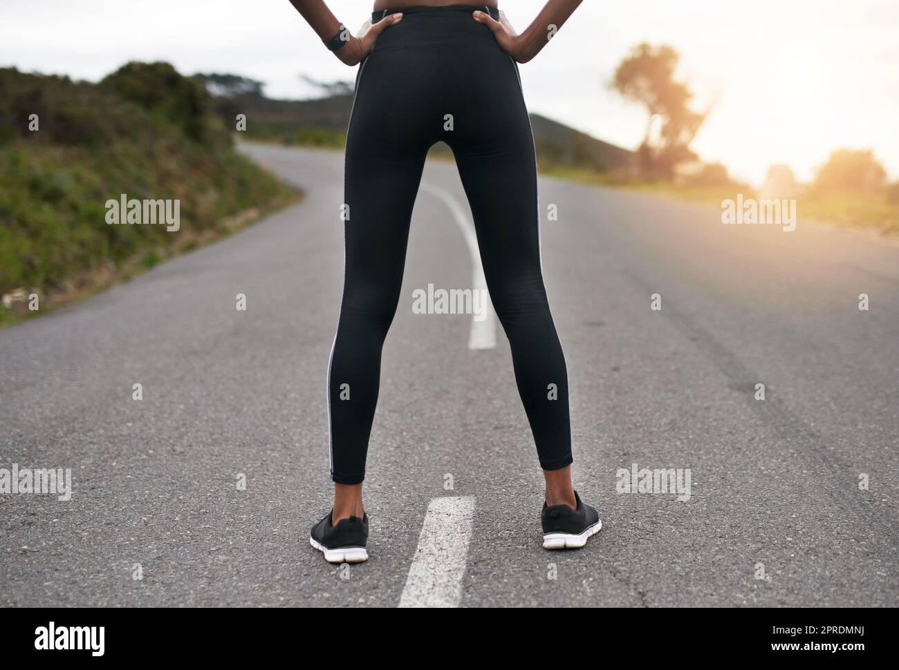 Die Straße weniger zurückgelegt. Rückansicht einer nicht erkennbaren jungen Sportlerin, die mit ihren Händen auf ihren Hüften draußen steht. Stockfoto