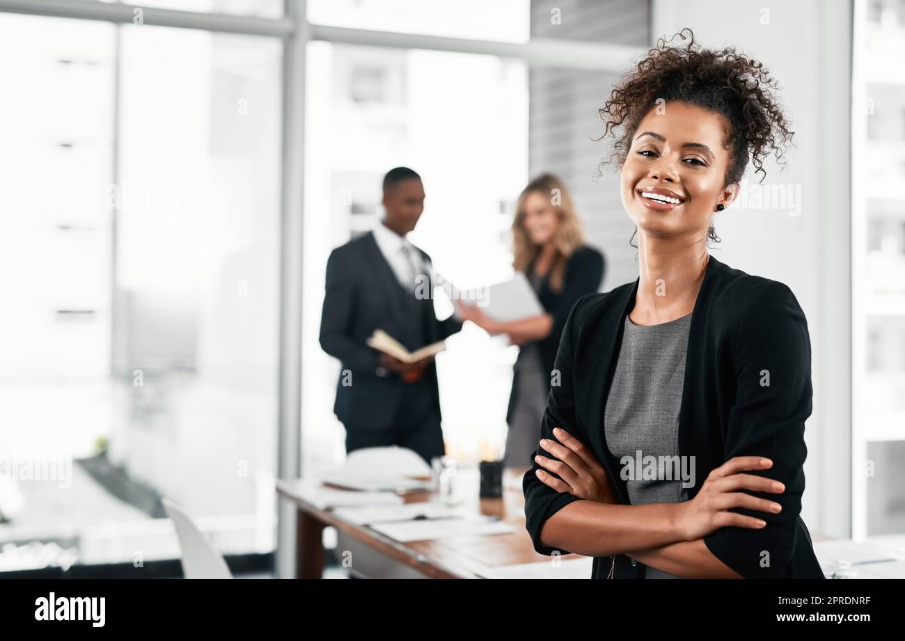 Ich freue mich, dass meine Karriere so positiv voranschreitet. Porträt einer jungen Geschäftsfrau, die in einem Büro mit ihren Kollegen im Hintergrund steht. Stockfoto