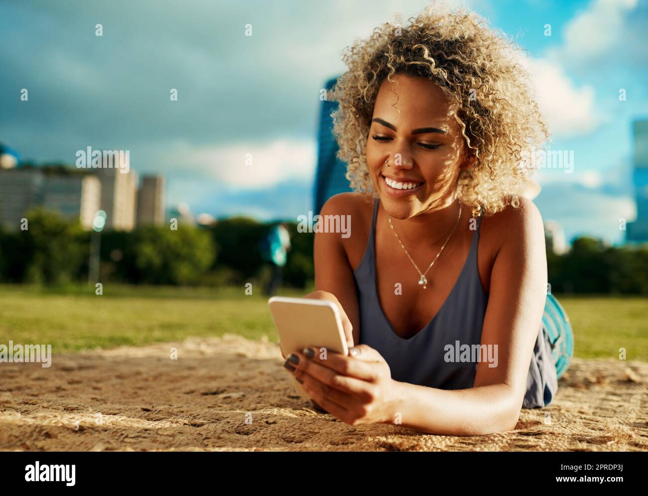 Will mich im Park begleiten. Eine attraktive junge Frau, die ein Smartphone benutzt, während sie sich in einem Park hinlegt. Stockfoto