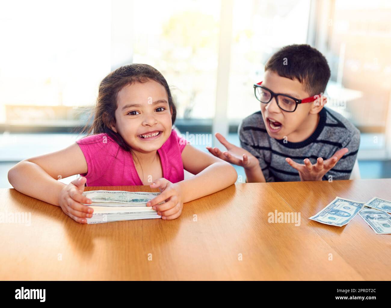 Sie gewinnt immer die Wetten zwischen ihnen. Ein entzückendes kleines Mädchen hält einen Stapel Geld, während ihr Bruder zu Hause ungläubig schreit. Stockfoto