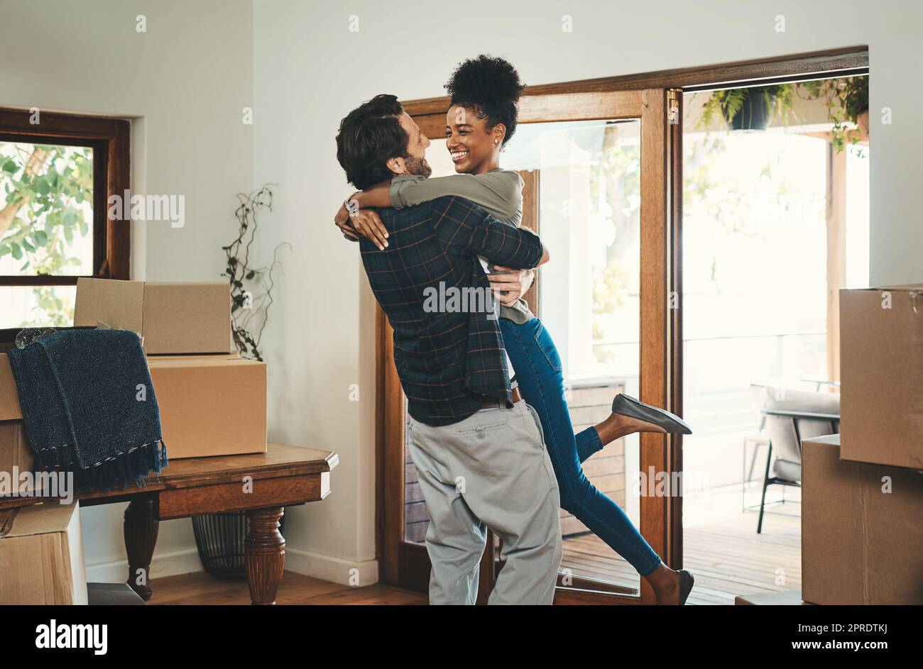 Hausbesitzer-Paar feiert, umarmt und jubelt in einem neuen Haus als Immobilieninvestoren, -Käufer und -Besitzer. Lustiger Mann und Frau aus verschiedenen Rassen, die aufgeregt, glücklich und fröhlich aussehen, während sie sich an Boxen umarmen Stockfoto