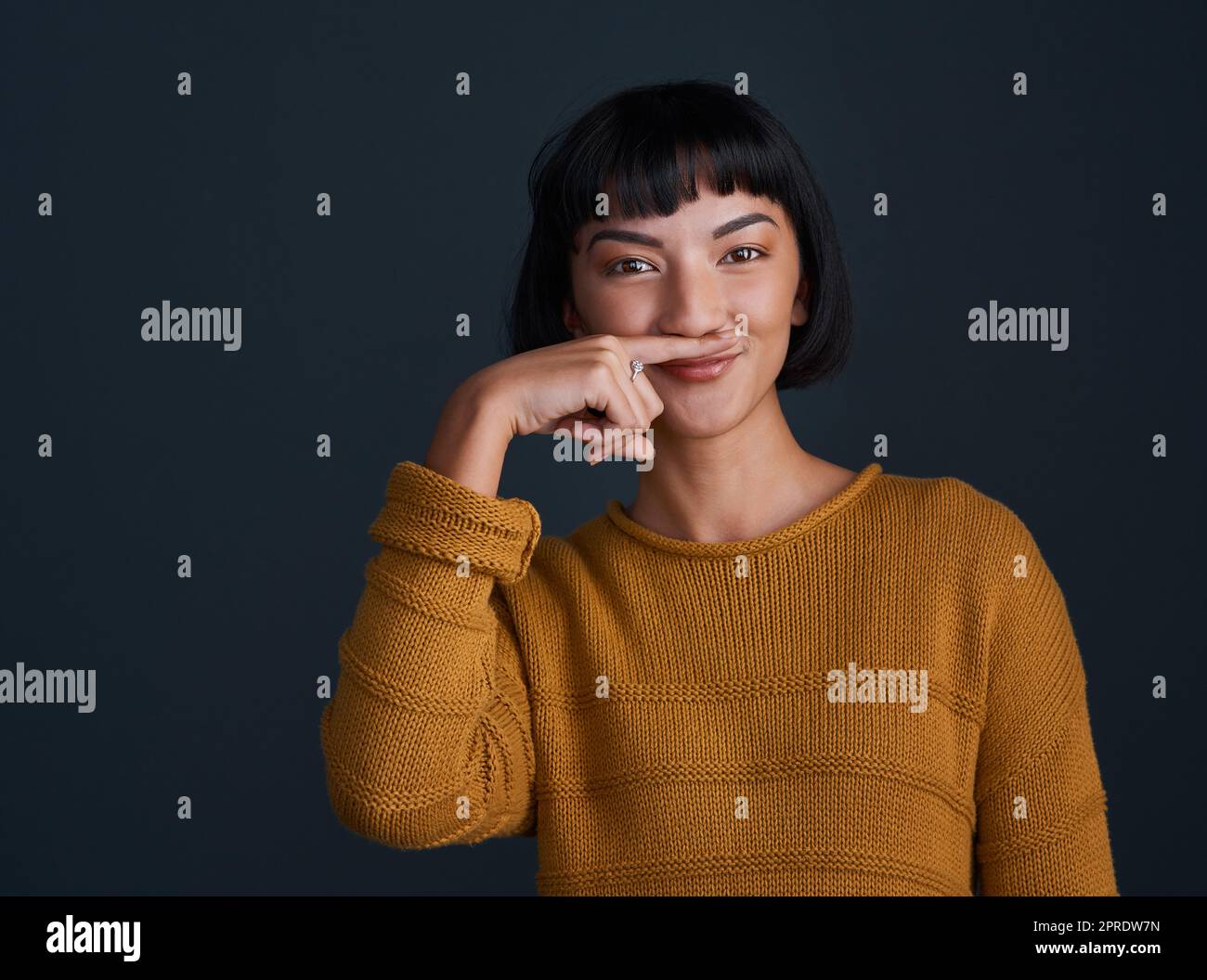 Was ist das Leben ohne ein bisschen Sillness. Studioaufnahme einer attraktiven jungen Frau, die mit ihrem Finger einen Schnurrbart vor einem dunklen Hintergrund macht. Stockfoto