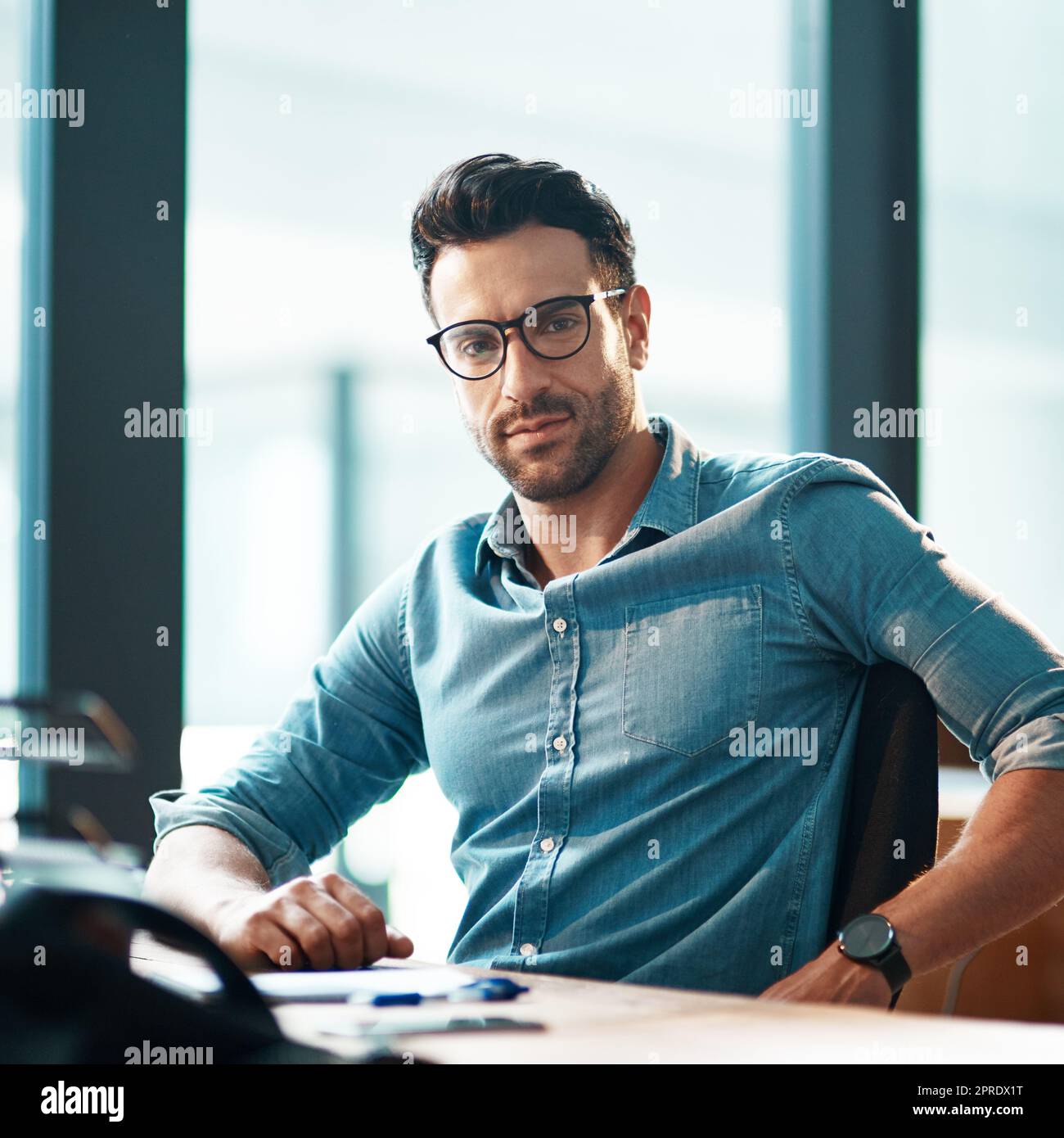 Junger, selbstbewusster und professioneller Geschäftsmann mit Brille, der an seinem Schreibtisch sitzt und in einem modernen Büroporträt arbeitet. Ein ungezwungener und seriöser Geschäftsmann in der Finanzabteilung Stockfoto