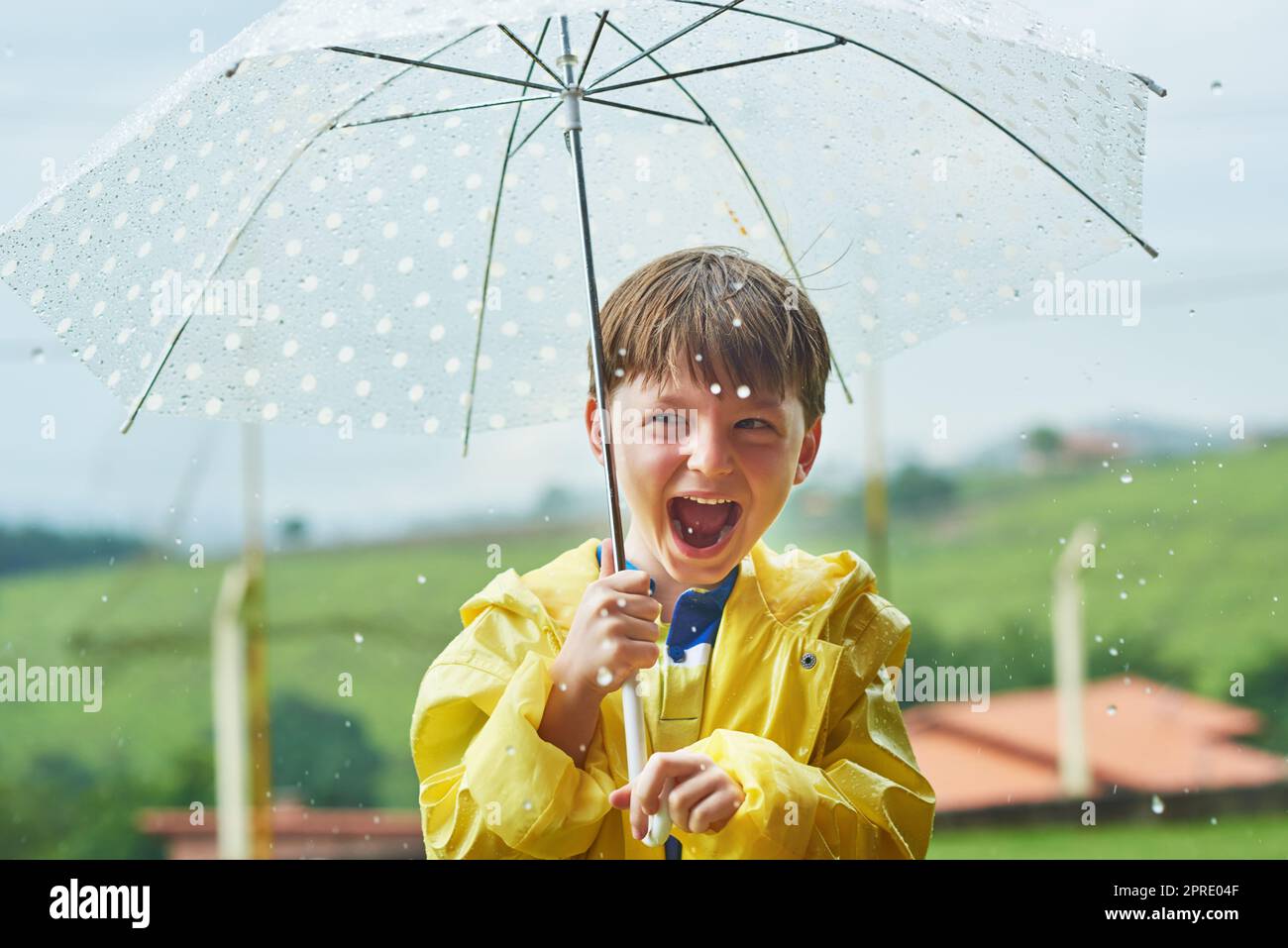 Die Regenzeit ist seine beliebteste Jahreszeit. Porträt eines fröhlichen kleinen Jungen, der an einem regnerischen Tag mit einem Regenschirm draußen steht. Stockfoto
