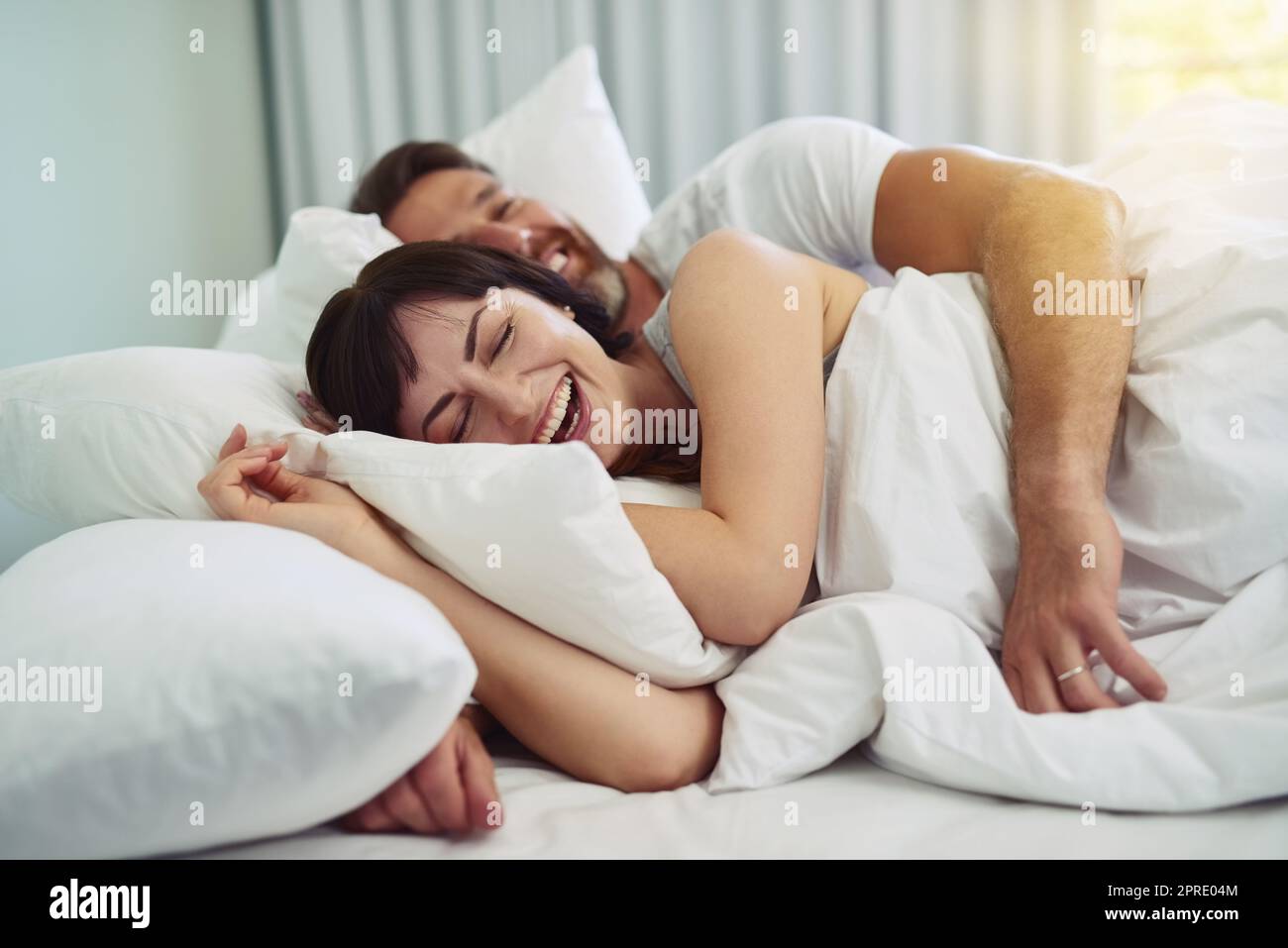 Sollten wir etwas länger im Bett bleiben? Ein entspanntes junges Paar, das versucht, in den Morgenstunden in den Armen des anderen im Bett zu schlafen. Stockfoto