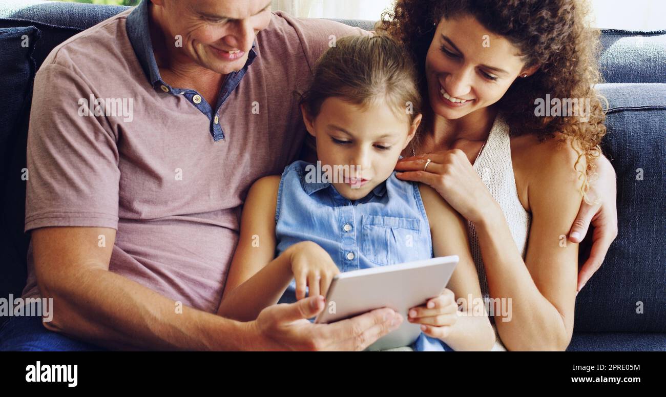 Im digitalen Zeitalter ist die Familienzeit die Bildschirmzeit. Ein süßes kleines Mädchen, das mit ihrer Mutter und ihrem Vater zu Hause auf dem Sofa ein digitales Tablet benutzt. Stockfoto