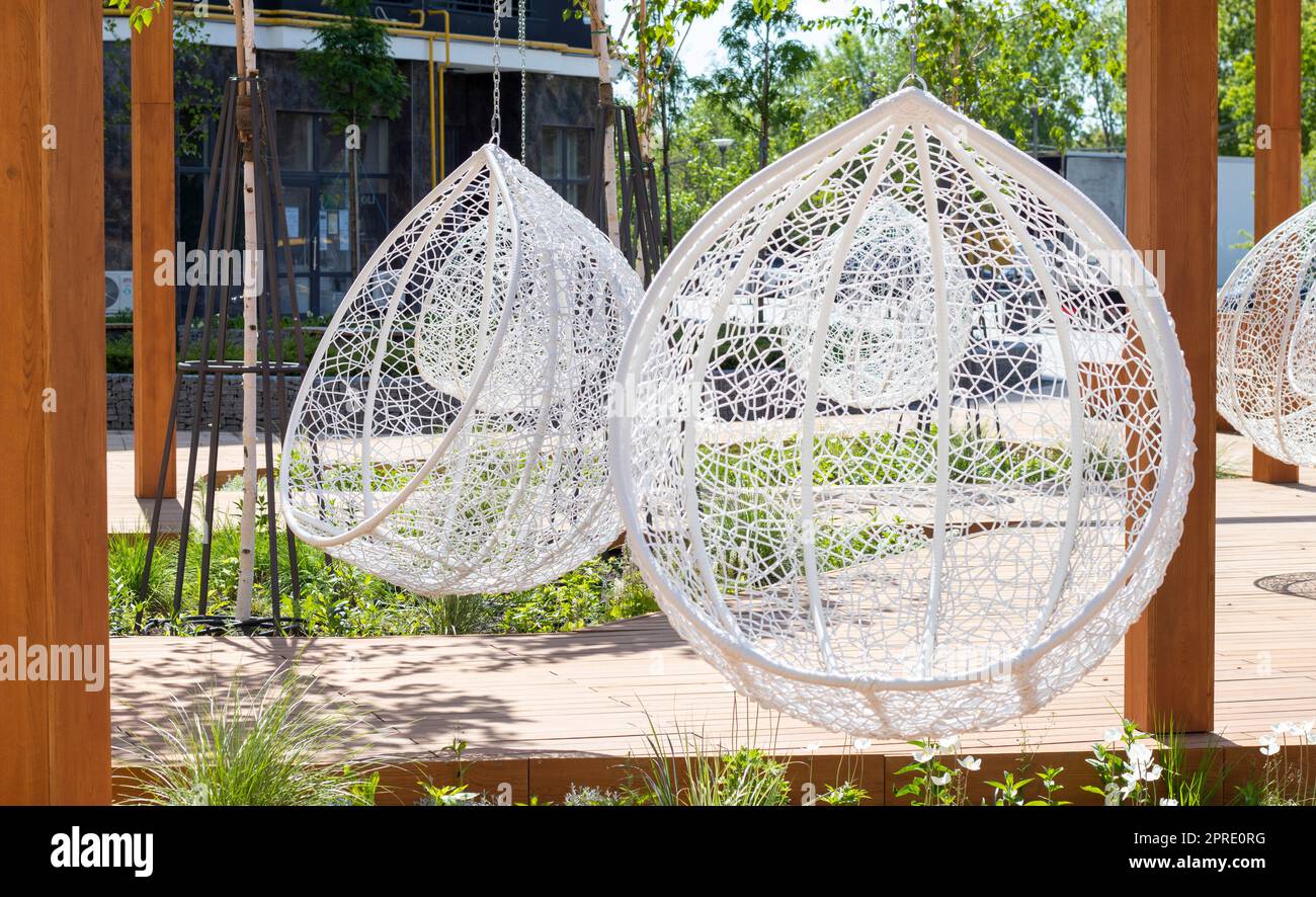 Ein gemütlicher Sitzbereich im Hof eines modernen Wohnkomplexes mit Holzbänken, weißen Korbstühlen, Girlanden und einem wunderschönen Landschaftsdesign mit Blumen und Rasen. Stockfoto