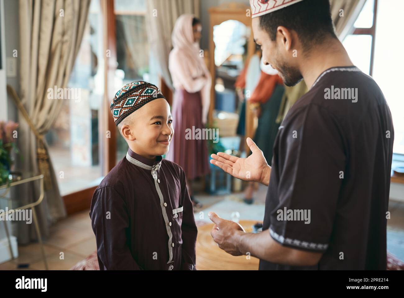 Muslimischer Vater und Sohn, die sich während eines religiösen Feiertages zu Hause im Wohnzimmer unterhalten und sich anfreunden. Süßer, lächelnder und glücklicher islamischer Junge, der Ratschläge von seinem Vater hört, während er ein traditionelles Outfit trägt Stockfoto