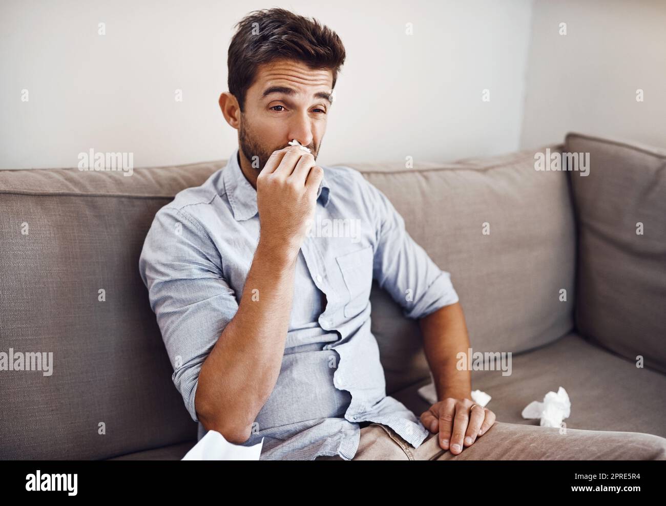 Eine verstopfte Nase zu haben ist so ärgerlich. Ein hübscher junger Mann bläst seine Nase mit einem Gewebe, während er sich auf einem Sofa zu Hause entspannt. Stockfoto