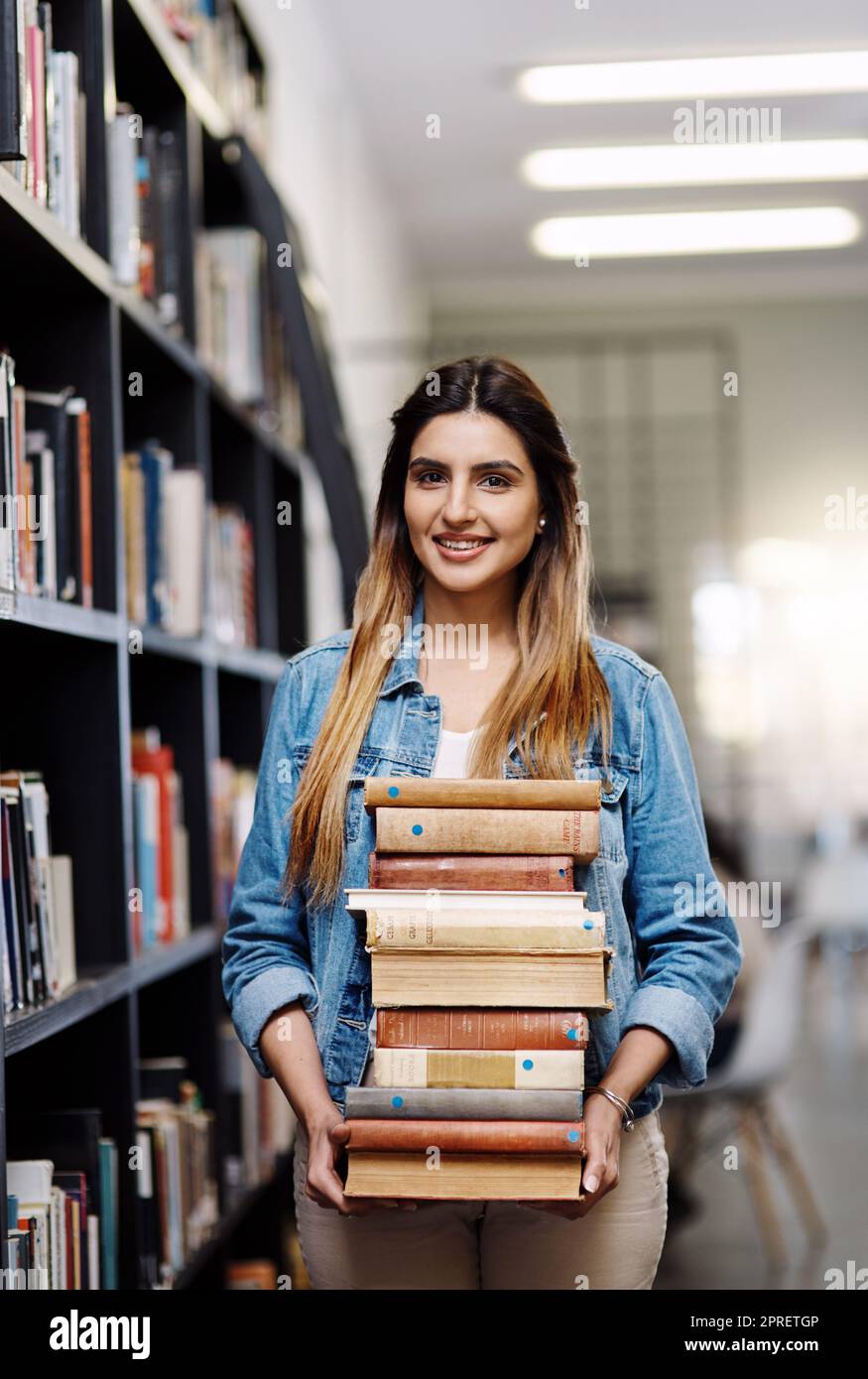 Das sind alle Bücher, die dieses akademische Jahr nicht brauchen. Porträt einer glücklichen jungen Frau, die Bücher in einer Bibliothek am College trägt. Stockfoto