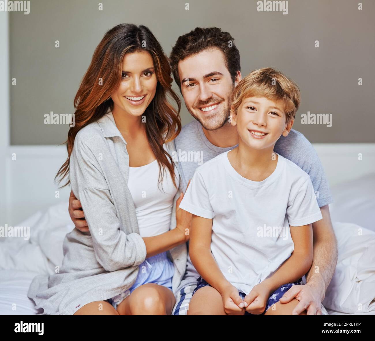 Fröhliche, glückliche Familie zu jeder Tageszeit. Porträt einer fröhlichen jungen Familie, die auf dem Bett saß, während sie sich gegenseitig festhängte und morgens zu Hause in die Kamera starrte. Stockfoto