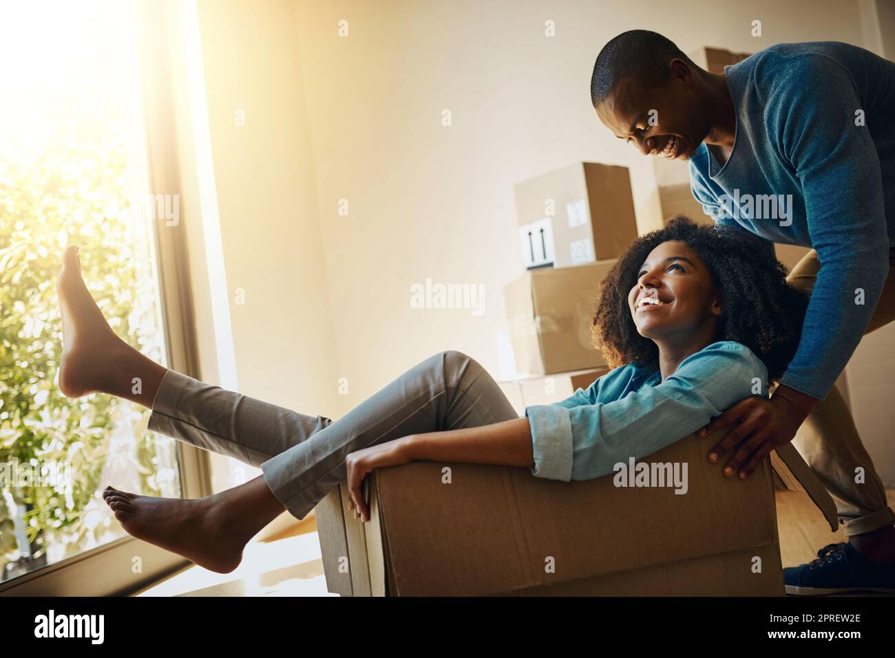 Wir wissen, wie man Spaß hat, egal in welcher Situation. Ein fröhlicher junger Mann schob seine Freundin während des Umzugstages in einem Karton zu Hause herum. Stockfoto