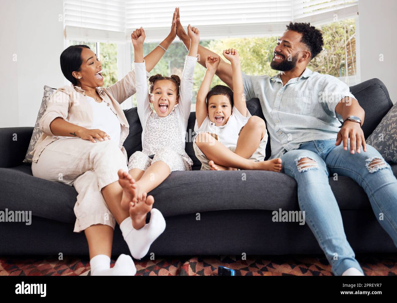 Glückliche Eltern und aufgeregte Kinder feiern auf dem Sofa, während sie fernsehen. Spaß mit der Familie, Entspannung zu Hause und Freundschaft. Mutter und Vater High Five, Kinder jubeln für den Sieg der Sportmannschaft. Stockfoto