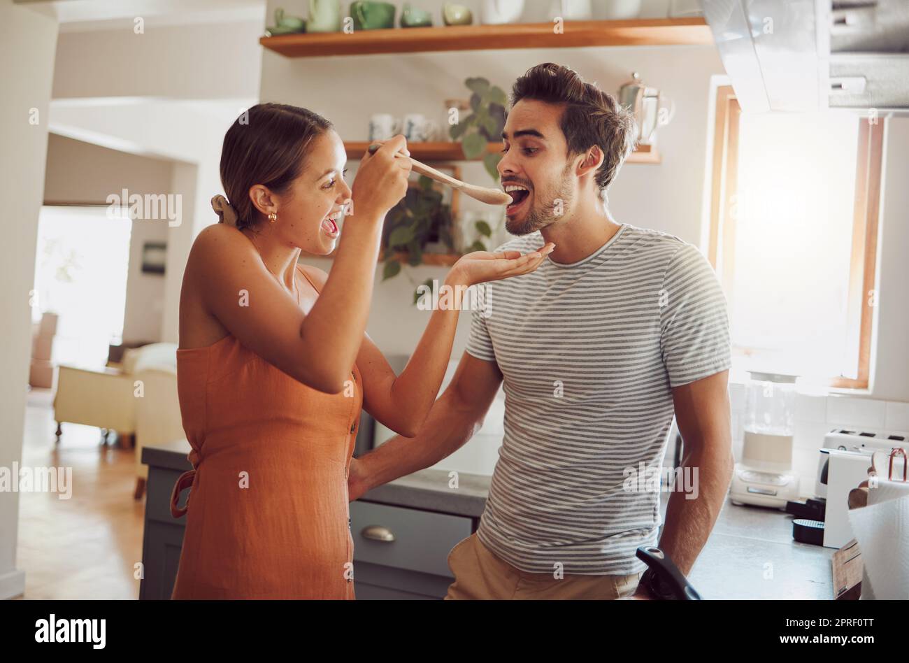 Spaß, Essen und ein junges Paar kochen in einer Küche zu Hause, sich verbinden, während sie verspielt sind und glücklich aussehen. Mann und Frau verkosten und flirten, kochen und machen zusammen Spaß Stockfoto