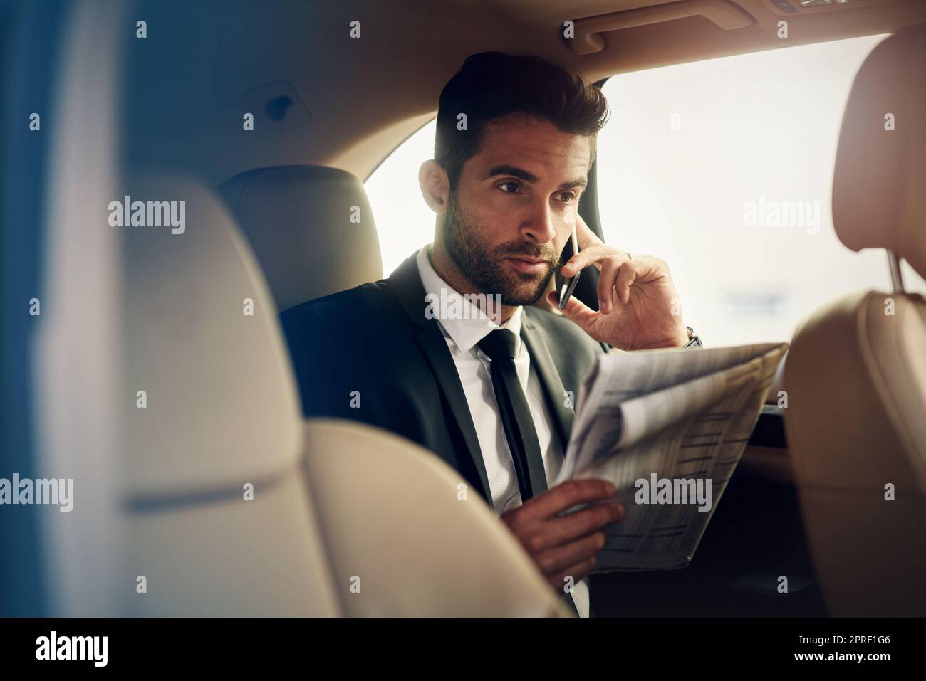 Überprüfen der Zahlen vor der Arbeit. Ein selbstbewusster junger Geschäftsmann, der am Telefon telefoniert und auf dem Rücksitz eines Autos eine Zeitung anschaut. Stockfoto