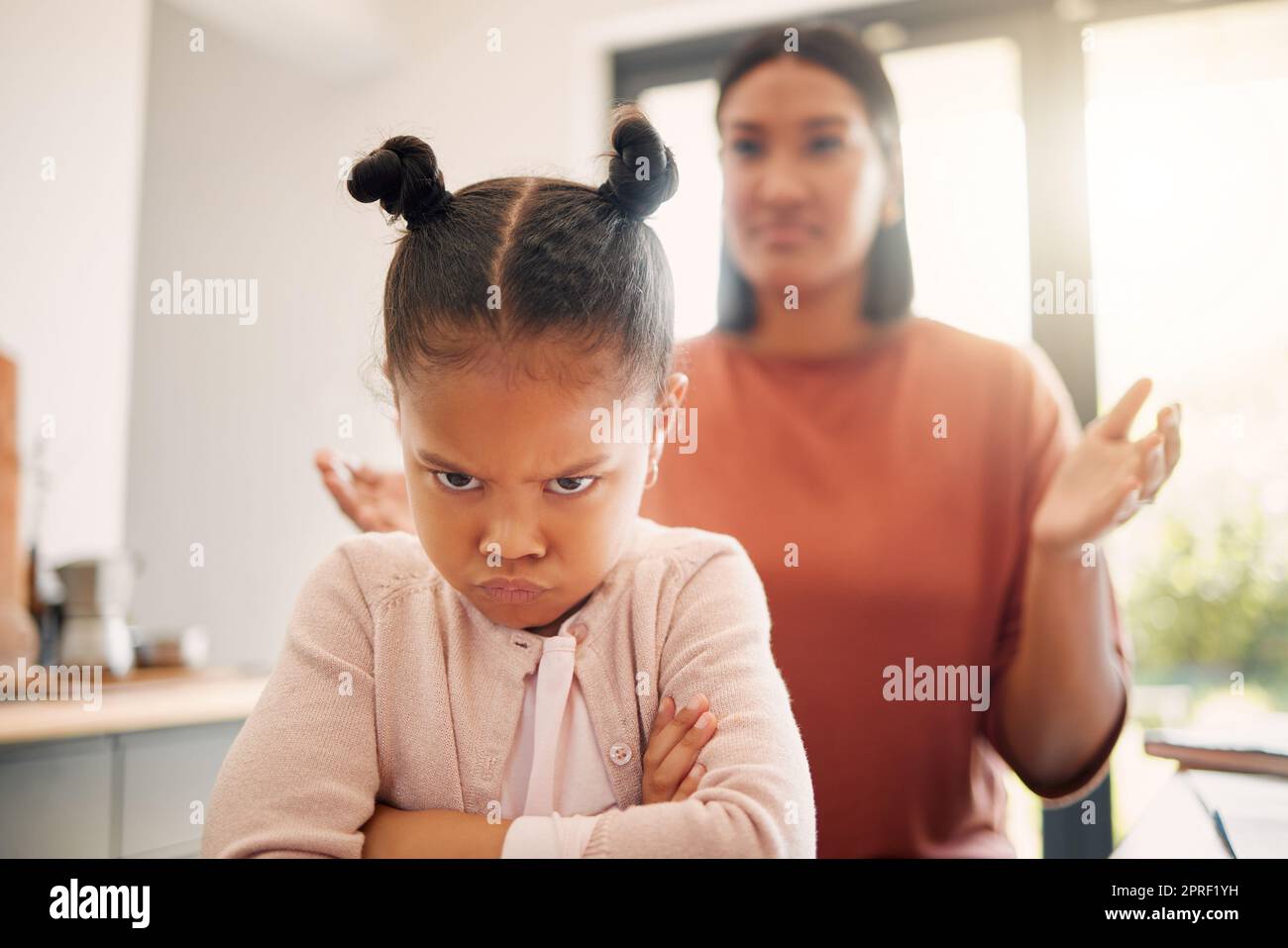 Wütendes, kleines Mädchen, unglücklich und aufgebracht nach dem Kampf oder von der Mutter geschimpft zu werden, die Stirnrunzeln vor Einstellung und verschränkten Armen. Unartiges Kind, das beleidigt aussieht, mit gestressten alleinerziehenden Eltern im Hintergrund. Stockfoto