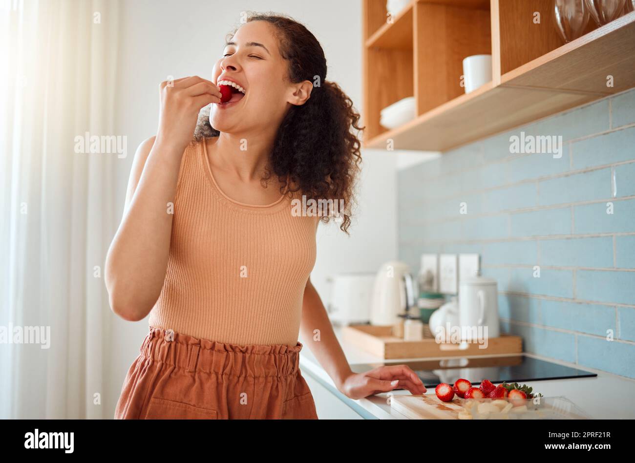 Essen, Zerkleinern oder Zubereiten von Obstsalat mit Erdbeeren, Bananen oder gesunden Zutaten in der Küche zu Hause. Lächelnde oder glückliche Frau, die frische Snacks kocht, beißt und verkostet oder das Frühstück zubereitet Stockfoto