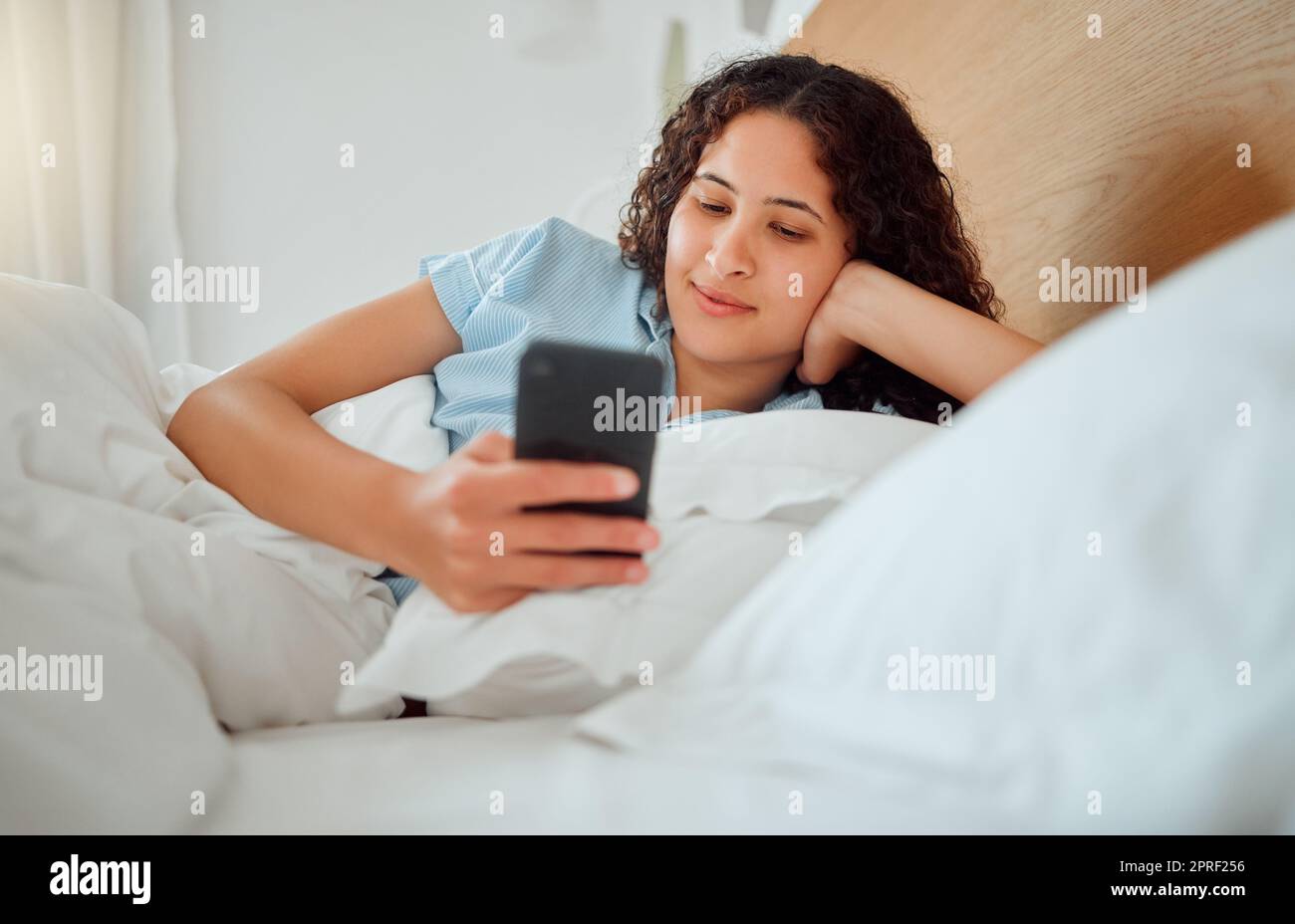 Eine Frau, die SMS liest, Nachrichten abruft und das Telefon hält, während sie sms liest und morgens wach in ihrem Bett liegt. Glückliche, zufriedene und lächelnde Frau, die ein Spiel spielt, im Internet oder in sozialen Medien surft Stockfoto