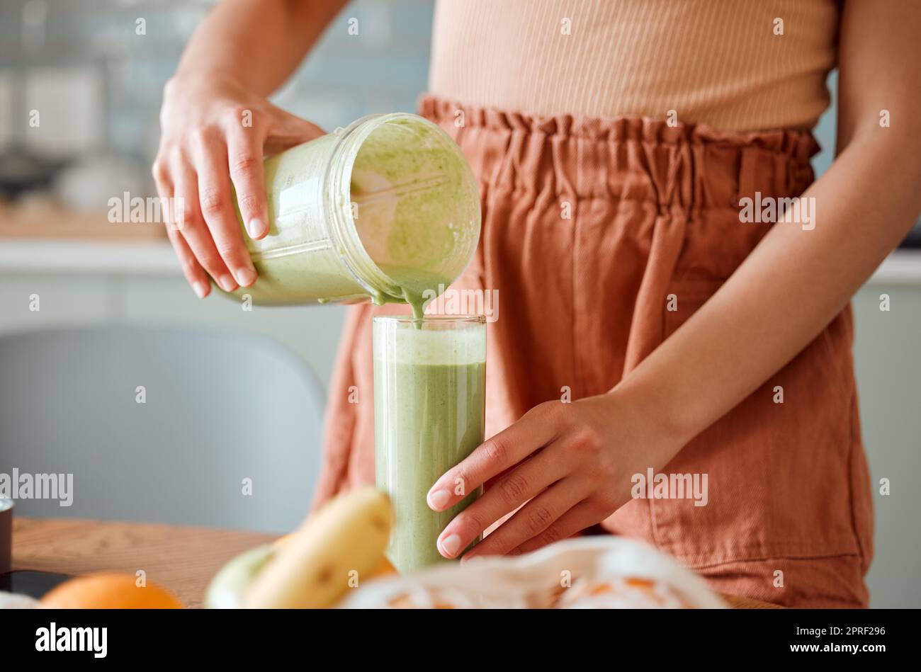 Frau, die einen gesunden Smoothie in ein Glas aus einem Mixbecher auf einer Theke zur Entgiftung eingießt. Eine Frau, die in ihrer Küche frischen grünen Fruchtsaft mit Gemüse und Verbrauchsmaterialien für einen gesunden Lebensstil herstellt. Stockfoto