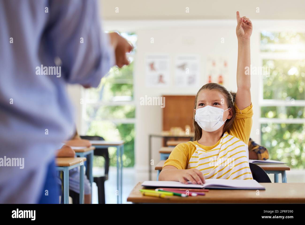 Kluger Schüler mit covid Gesichtsmaske, der Lehrer Frage über Corona-Virus-Pandemie in einem Klassenzimmer oder Grundschule stellen. Kleines Mädchen hebt Hand, um Gesundheitsthemen im Unterricht zu beantworten Stockfoto