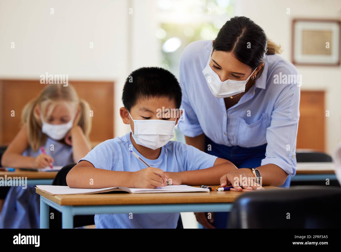 Covid, Bildung und Lernen mit einem Lehrer, der eine Maske trägt und einem männlichen Schüler während der Schule hilft. Ein Junge, der in einem Klassenzimmer lernt, mit Hilfe eines Pädagogen, während er an seinem Schreibtisch sitzt. Stockfoto