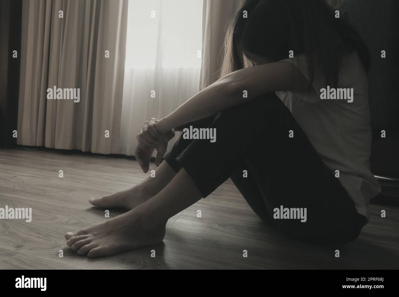 Depressive und gestresste Frau, die auf dem Boden neben den Vorhängen im Schlafzimmer saß. Traurige Frau, die im dunklen Raum sitzt. Mädchen mit psychischen Problemen. Unglückliches Leben. Weltkonzept für den Tag der psychischen Gesundheit. Stockfoto