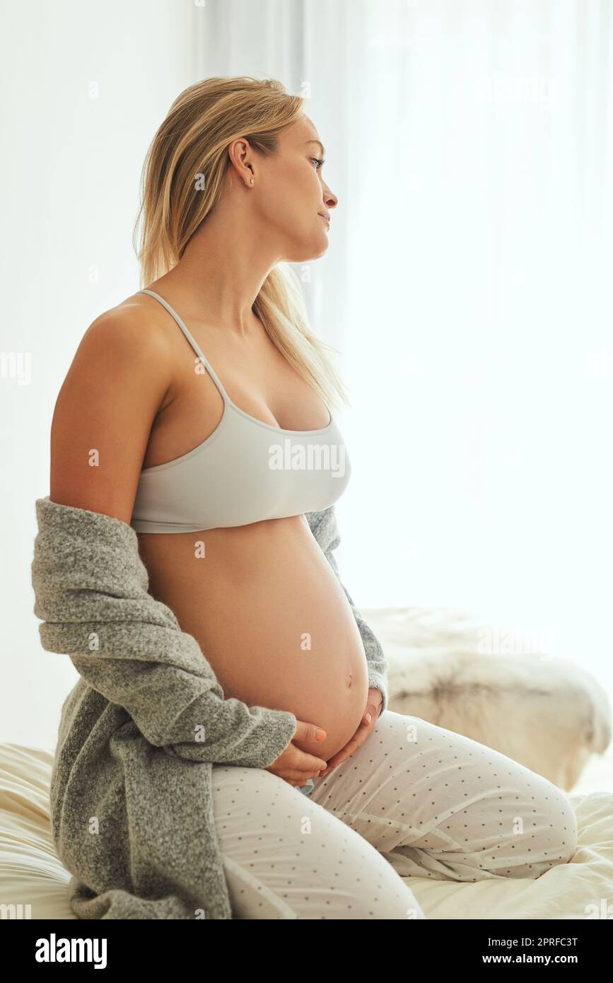 Die Schwangerschaft ist eine erstaunliche Reise. Eine Schwangere berührt ihren Bauch, während sie auf ihrem Bett sitzt. Stockfoto