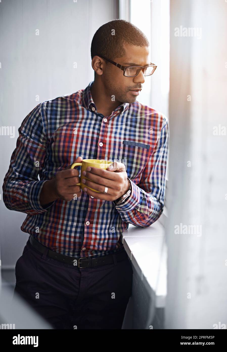 Ein Geschäftsmann, der eine Pause macht, während er aus dem Fenster schaut. Ein afroamerikanischer Mann, der sich drinnen entspannen kann, indem er sich die Aktivitäten im Freien anschaut. Ein Büroangestellter beobachtet das Stadtleben unter dem Gebäude. Stockfoto