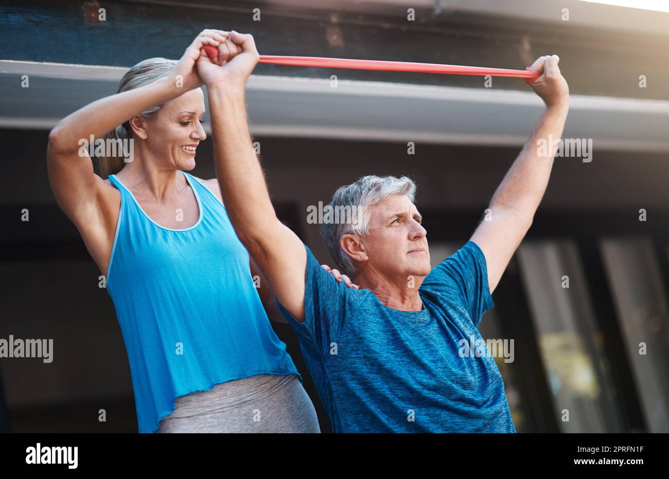 Sie gibt mir immer diesen zusätzlichen Schub. Ein reifer Mann streckt seine Armmuskeln mit einem Gummiband, während er von seiner Frau unterstützt wird. Stockfoto